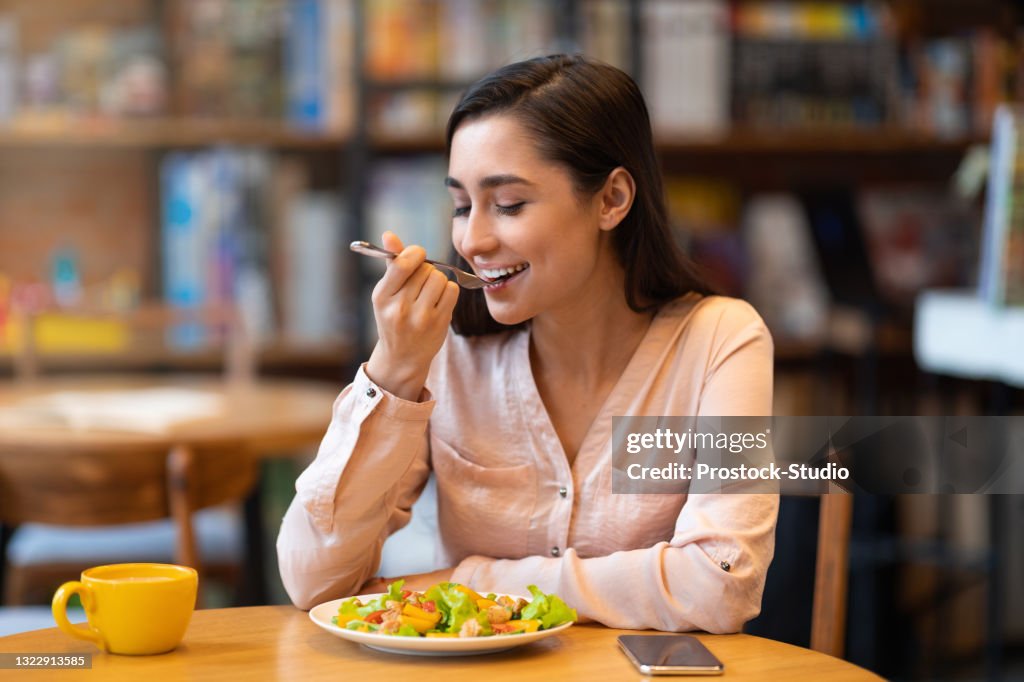 Mulher latina feliz almoçando no café, desfrutando de salada deliciosa com olhos fechados e bebendo bebida quente