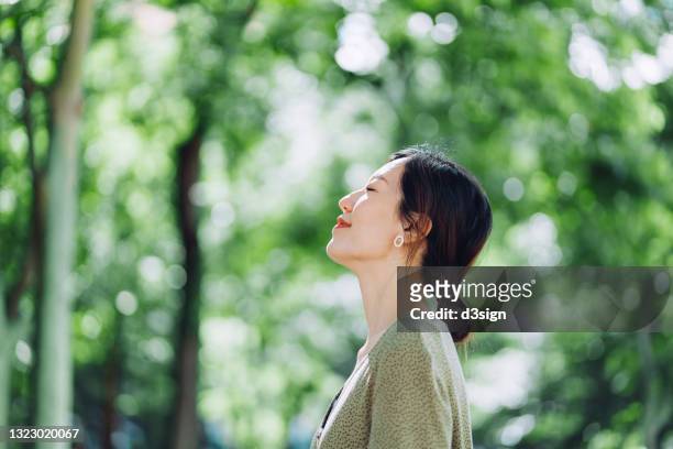 beautiful young asian woman meditating in the nature with her eyes closed, setting herself free and feeling relieved. enjoying fresh air and breathing in the calmness with head up against sunlight in the morning. freedom in nature. connection with nature - vivere semplicemente foto e immagini stock