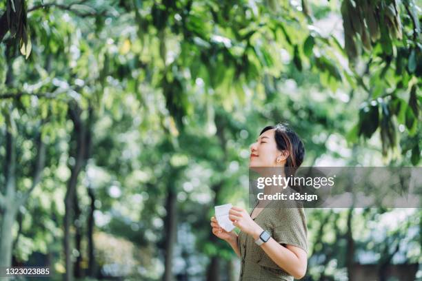 beautiful young asian woman taking off protective face mask, setting herself free and feeling relieved. head up. enjoying some fresh air with her eyes closed while relaxing in park. hope for an end of pandemic concept - air pollution mask stockfoto's en -beelden