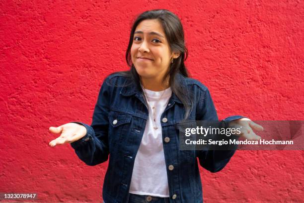 young latino woman shrugging with her hands to her sides, red background - stupid stock-fotos und bilder