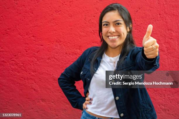 young latino woman looking at the camera and giving a thumbs up, red background - fresh deals ストックフォトと画像