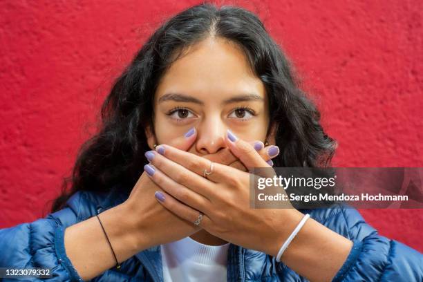 young latino woman looking at the camera and covering her mouth, speak no evil, close up, red background - hand voor de mond stockfoto's en -beelden