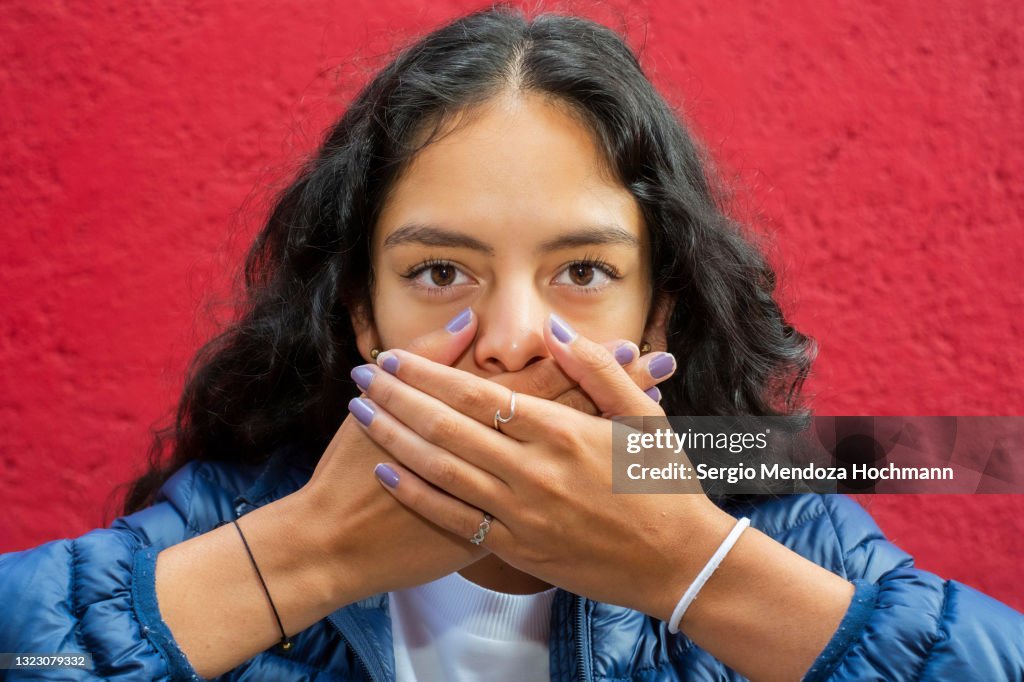 Young Latino woman looking at the camera and covering her mouth, SPEAK NO EVIL, close up, red background