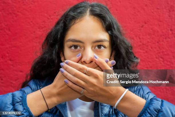 young latino woman looking at the camera and covering her mouth, speak no evil, close up, red background - hands covering mouth stock pictures, royalty-free photos & images
