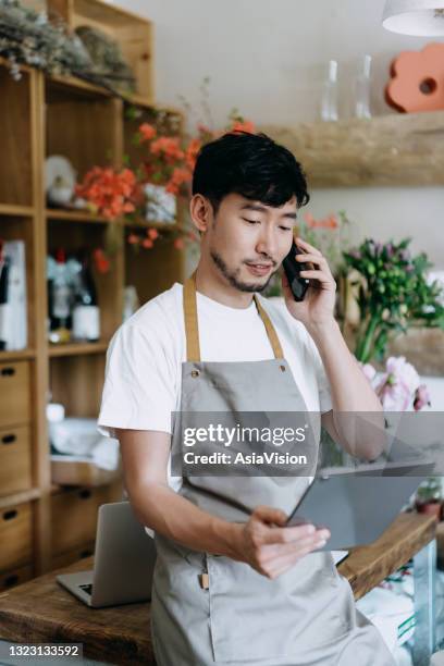 junge asiatische männliche floristin, inhaber in klein unternehmenden blumenladen, sprechen auf dem smartphone, während die arbeit auf digitalen tablet am arbeitsplatz. aktien prüfen, kundenbestellungen entgegennehmen, produkte online verkaufen. täglic - asien e commerce stock-fotos und bilder