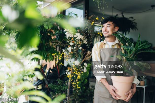 zekere jonge aziatische mannelijke bloemist, eigenaar van kleine bedrijfsbloemwinkel. het houden van potplant buiten zijn werkplaats. hij kijkt weg met een glimlach. geniet van zijn werk om bij de bloemen te zijn. klein bedrijfsconcept - shop stockfoto's en -beelden