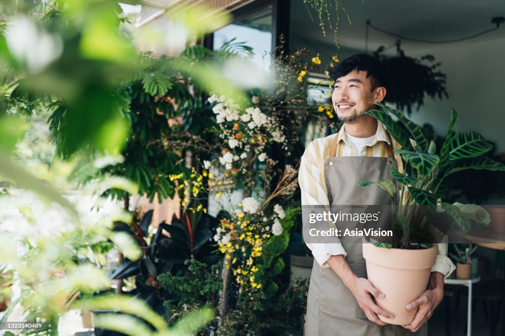 自信的年輕亞洲男花店,小企業花店老闆。在他的工作場所外拿著盆栽植物。他面帶微笑地望向遠方。享受他的工作,與花。小型企業概念