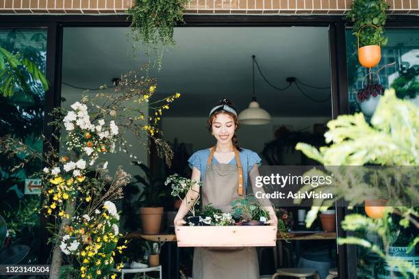 cheerful young asian female florist, owner of small business flower shop. holding a wooden crate full of flowers and plants outside her workplace. she is smiling while looking at the plants. enjoying her job to be with the flowers. small business concept - full responsibility stock pictures, royalty-free photos & images