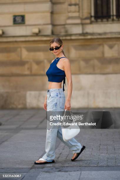 Xenia Adonts wears sunglasses, golden earrings, a beaded necklace with a blue mini teddy bear, a blue off-shoulder back-nude cropped tank top, pale...
