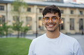 Young successful Indian man wearing stylish eyeglasses, standing on the street. Handsome asian model posing for pictures, looking at camera, smiling