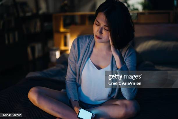 stressed young asian woman with smartphone sitting on bed in bedroom with hand in head, encountering sleeping problems and insomnia at night - asian woman pregnant stock pictures, royalty-free photos & images