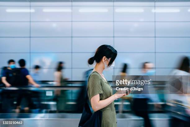 side profile of young asian woman with protective face mask using smartphone surrounded by commuters rushing by in subway station during office peak hours in the city - crowd of people walking stock pictures, royalty-free photos & images