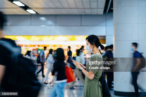 side profile of young asian woman with protective face mask using smartphone surrounded by commuters rushing by in subway station during office peak hours in the city - crowd masks stock pictures, royalty-free photos & images