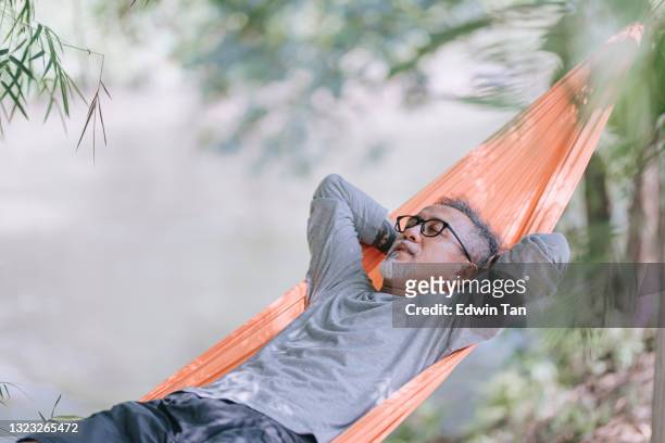 asian chinese senior man enjoying his afternoon lying down at hammock at riverside under bamboo trees - hammock stockfoto's en -beelden