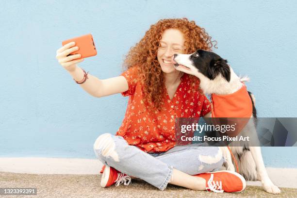 redheaded woman sitting on the ground with her dog taking a selfie against a blue wall while the dog kiss her - bandana fotografías e imágenes de stock