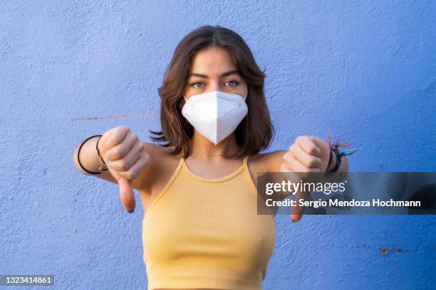 young latino woman wearing a face mask, looking at the camera and giving two thumbs down, blue background - funny face mask stock-fotos und bilder