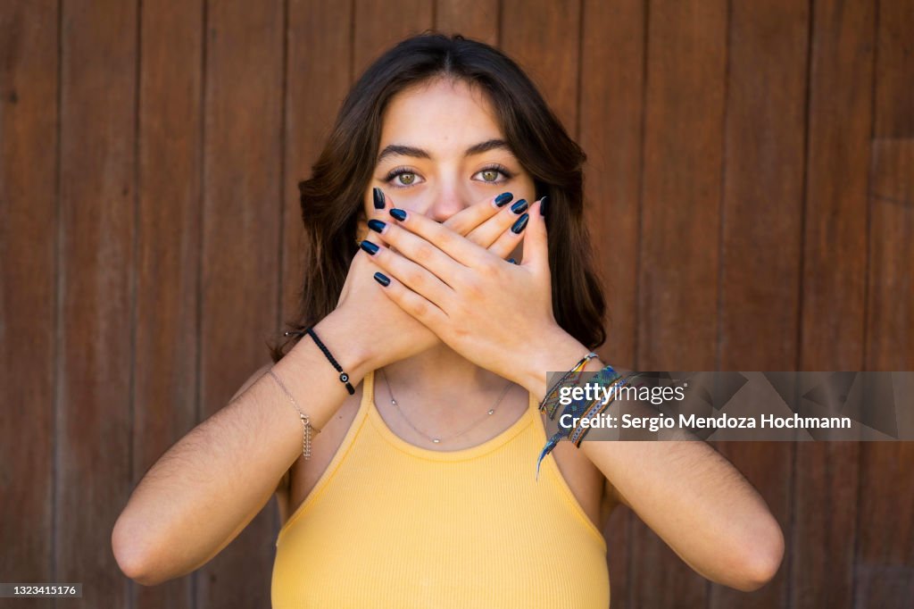 Young Latino woman looking at the camera and covering her mouth, SPEAK NO EVIL, brown wooden background