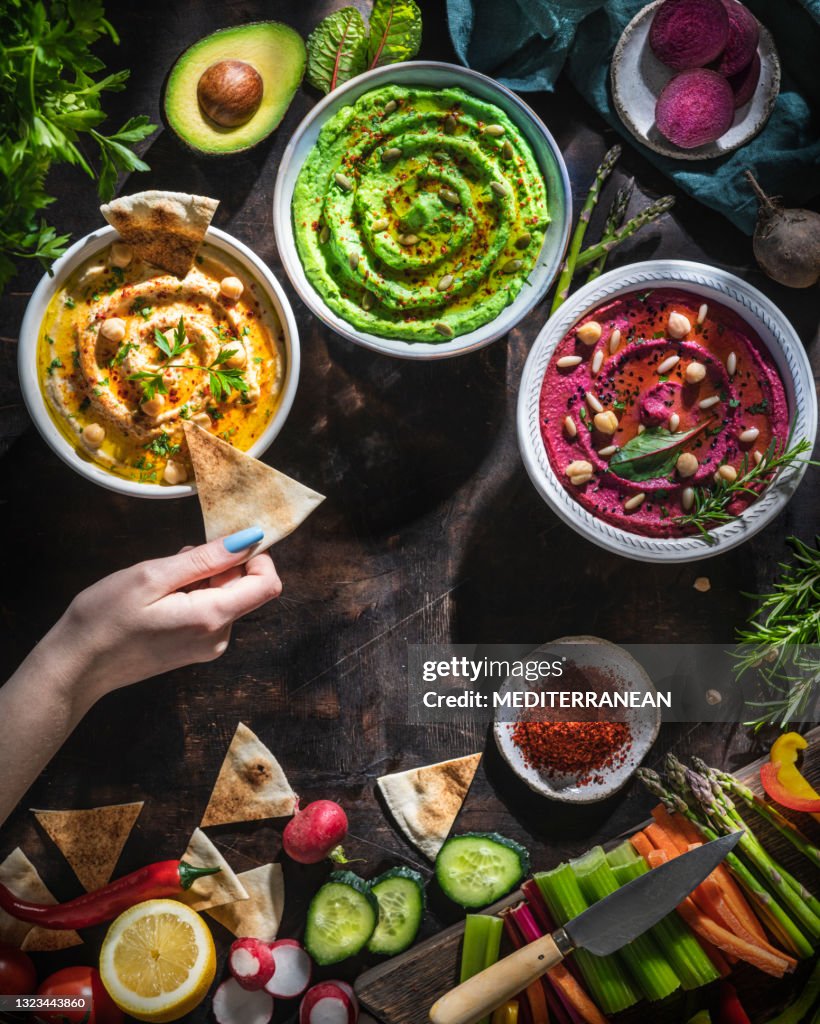 Hummus bowls of chickpeas, avocado and beetroot female hand dip with cut vegetables for dipping