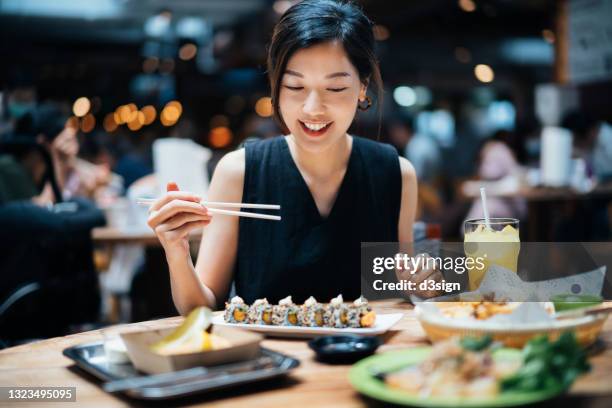 cheerful young asian woman enjoying scrumptious dishes and various cuisines from around the world in an exciting gourmet food hall. she cannot wait to eat all these tasty food. international cuisine and food culture. eating out lifestyle - eating sushi stock-fotos und bilder