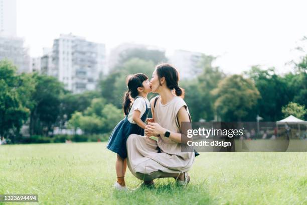 a loving young asian mother kissing and embracing her lovely little daughter in urban park, against beautiful sunlight. precious moment between mother and daughter. family lifestyle. family love and care. enjoying nature and summer days outdoor - chinese mothers day foto e immagini stock