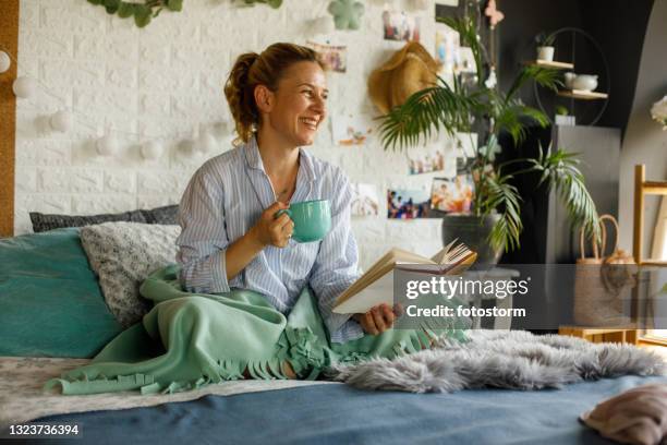 cheerful woman enjoying morning coffee in bed while reading a book - hygge stockfoto's en -beelden