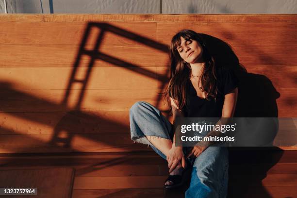 beautiful young woman enjoying the sunlight with her eyes closed while sitting on the wooden floor - moment of silence stockfoto's en -beelden