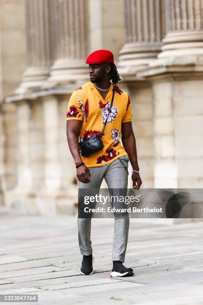 Cali Chiki wears a red beret hat, a silver and diamond chain necklace, a yellow mustard polo shirt white white / red and burgundy print pattern, a...