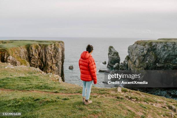 young woman relaxes at viewpoint on rocky coastline - puffer jacket stock pictures, royalty-free photos & images