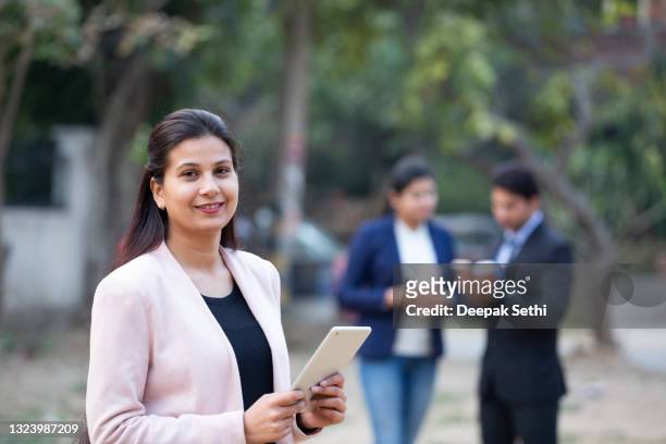 portrait of indian businesswoman using digital tablet, her colleagues communicating in background:- stock photo - indian corporate women background stockfoto's en -beelden