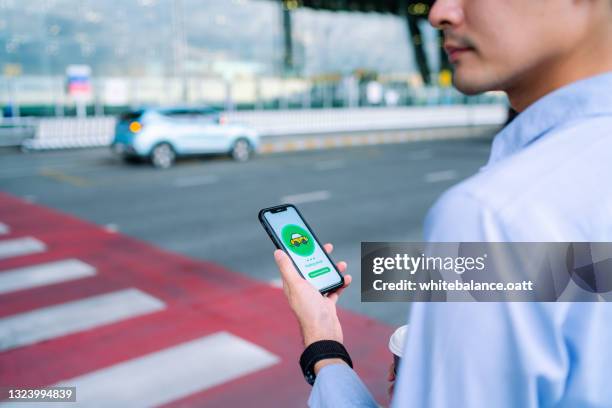 young asian man using mobile app device on smartphone to order a taxi pick up service by the urban road in downtown city street. speedy and trustworthy service. carsharing and business on the go themes - carsharing stock pictures, royalty-free photos & images