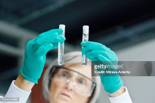 a senior caucasian female lab technician, looks at a test tube while working in a laboratory, wearing goggles, a white lab coat and green protective gloves. - lab closeups stock-fotos und bilder