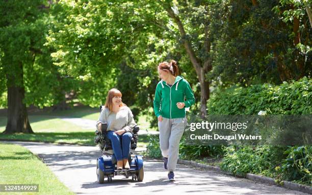 woman in wheelchair and friend running along side in park. - motorized wheelchair stock pictures, royalty-free photos & images