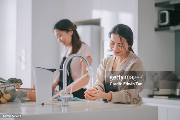 アジアの中国の女性の手洗浄は、ラップトップを使用してキッチンカウンターで週末に自宅で流水で流しでオレンジ色の果物を洗浄 - 浄水 ストックフォトと画像