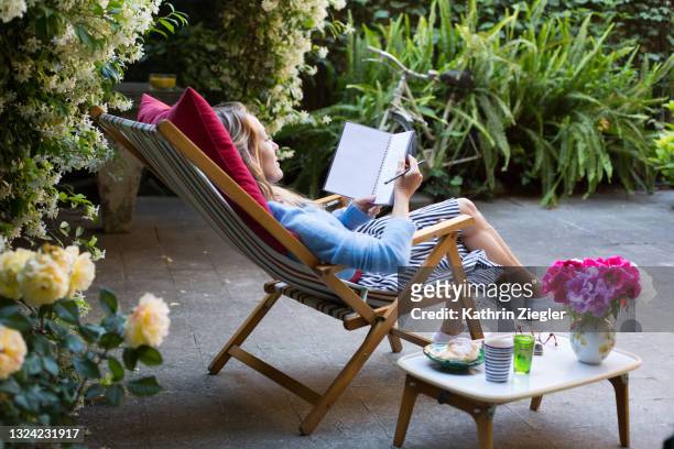 woman relaxing on deck chair in back yard, writing in notebook while having breakfast - backyard deck stockfoto's en -beelden