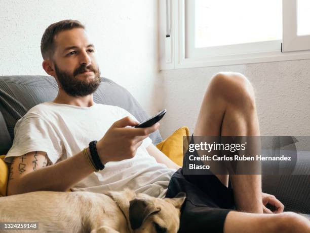 young man relaxing at home lying next to his dog while changing the tv channel. - alter tv stock pictures, royalty-free photos & images