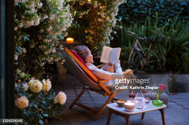 woman relaxing on deck chair in back yard, reading a book with her cat watching - leggere foto e immagini stock