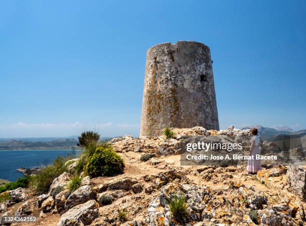 woman contemplating an ancient medieval watchtower on top of a mountain by the sea in majorca island. - puerto pollensa stock-fotos und bilder