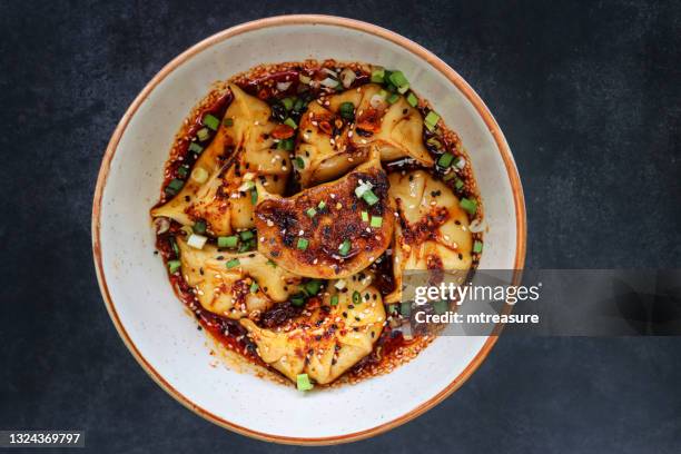 image of white bowl of momos (south asian dumplings), pan fried, white flour and water dough filled with chicken and mixed vegetables, drizzled with chilli oil and garnished with chopped chives and nigella seeds, black background, elevated view - dim sum meal stock pictures, royalty-free photos & images