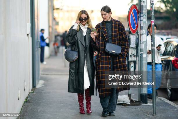 Guest wears black sunglasses, gold earrings, a white turtleneck sweatshirt, a long oversized black shiny leather trench coat, a long white shirt, a...