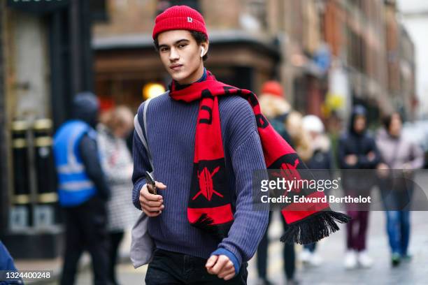Guest wears a red wool ribbed beanie, a blue woven wool pullover, a red with black inscription scarf, a blue ribbed turtleneck pullover, black denim...