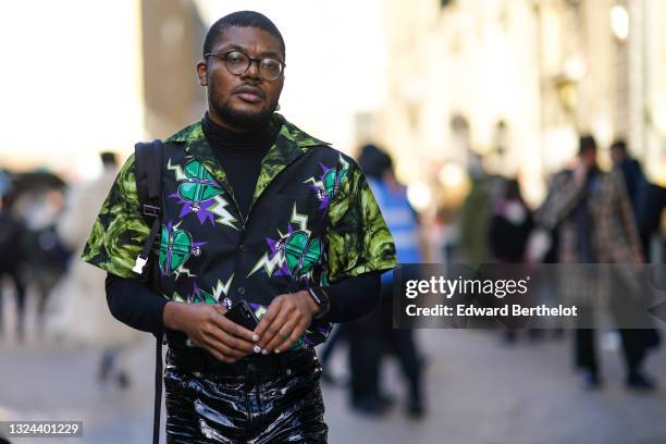 Guest wears black glasses, a black wool turtleneck pullover, a black and green tie and dye with green and purple heart pattern Prada short sleeves...