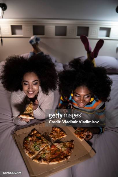 african women relaxing at home and eating pizza on bed - african family watching tv stockfoto's en -beelden