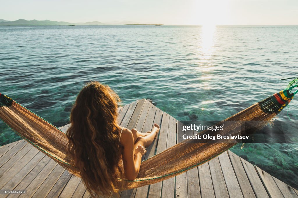 View from behind of woman awakening with ocean view. Wooden hotel terrace with hammock. Beautiful morning light
