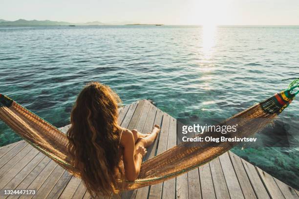view from behind of woman awakening with ocean view.  wooden hotel terrace with hammock. beautiful morning light - carefree stock pictures, royalty-free photos & images