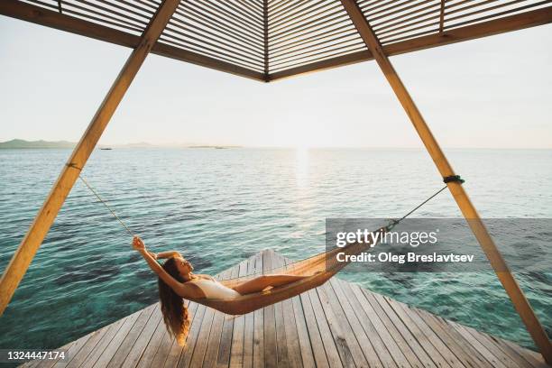 woman enjoying in hammock on wooden hotel terrace with ocean view. beautiful sunset light - beach glamour stock pictures, royalty-free photos & images