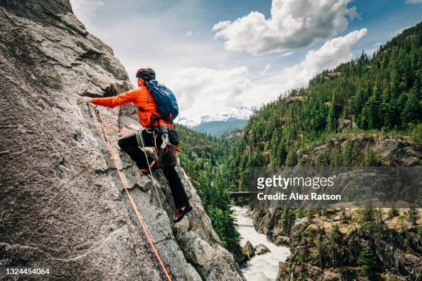 a male rock climber leads up a rock face high above a mountains river valley near whistler, bc - klettergarten stock-fotos und bilder