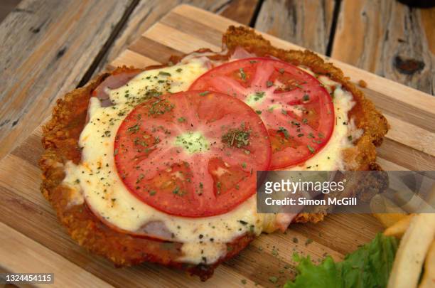 milanesa a la napolitana (thin slice of fried beef filet topped with tomato sauce, ham, mozzarella cheese and fresh tomato slices) on a wooden cutting board - milanese stockfoto's en -beelden