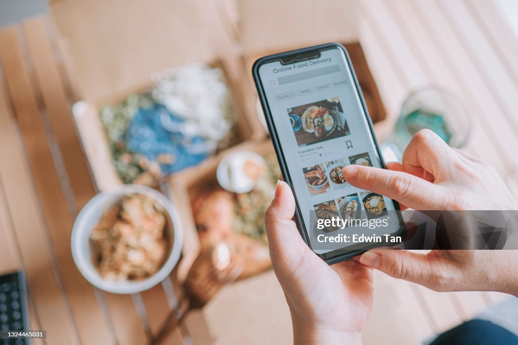 Hochwinkelansicht Nahaufnahme asiatische Frau mit Essen Lieferservice Bestellen Lebensmittel online mit mobilen App auf Smartphone im Wohnzimmer in einem gemütlichen Haus
