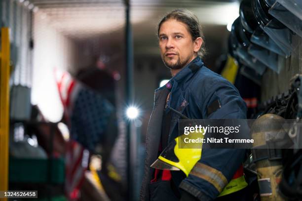 pride of male firefighter wearing fire protection suit and holding helmet while standing and looking away at fire station. - firefighter getting dressed stock pictures, royalty-free photos & images