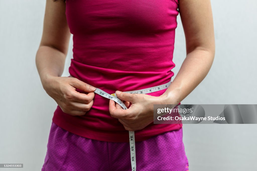 Midsection of adult slim woman in sportswear measuring waist circumference. Inch scale of measuring tape.